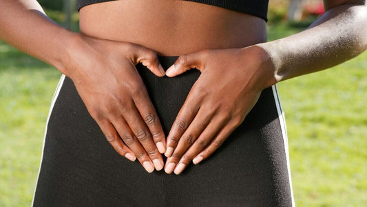 A young black woman's torso is shown, clothed in athletic gear that shows her midriff, which has her hands resting on it, forming a heart shape with her hands