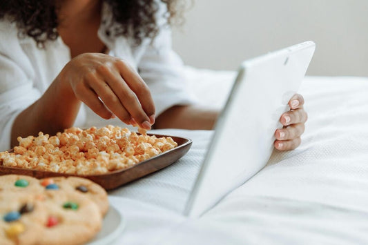 A young woman with her face out of shot lazily grazing on cookies an dpopciorn as she uses a laptop on a white bedspread.